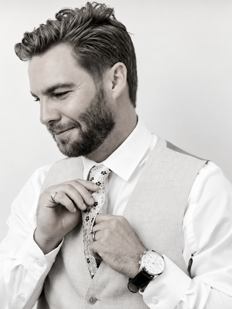 Daniel Jeffery in a white collared shirt and vest, adjusting his tie